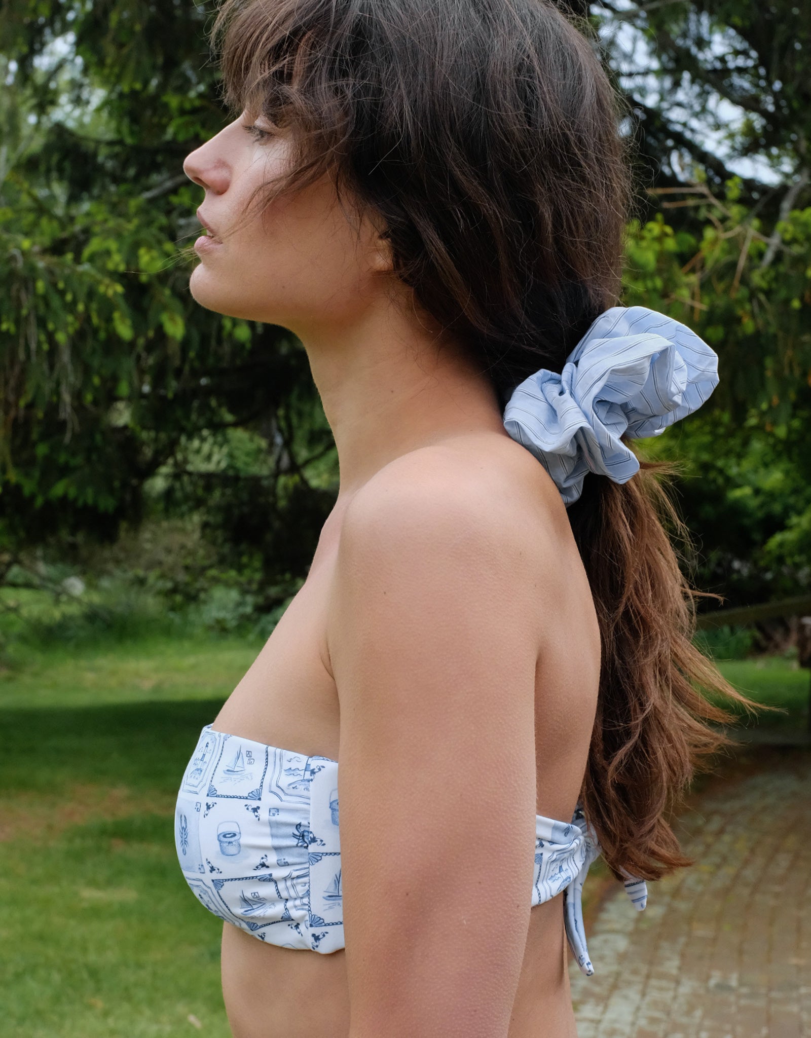 Woman wearing a strapless bikini with a blue and white pattern, standing outdoors with greenery in the background.
