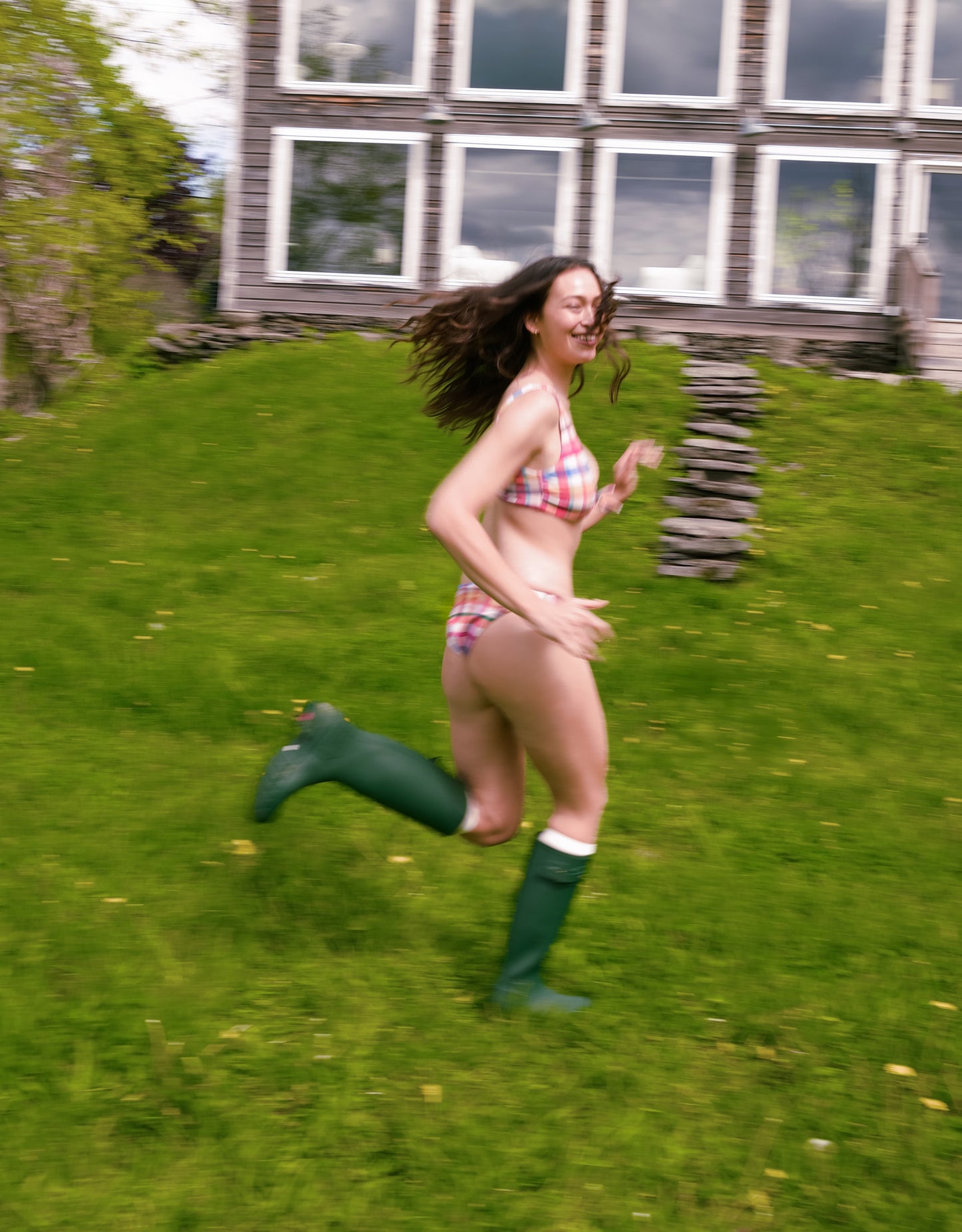 Person running on grass wearing a bikini and green rain boots with a house in the background.