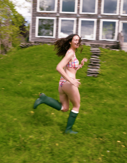 Person running on grass wearing a bikini and green rain boots with a house in the background.